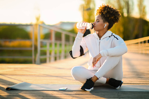 Dorstige jonge Afro-Amerikaanse vrouw drinkwater tijdens outdoor training, sportieve zwarte vrouw zittend op fitness mat en genietend van gezonde drank, rusten na training buiten, kopieerruimte - Foto, afbeelding