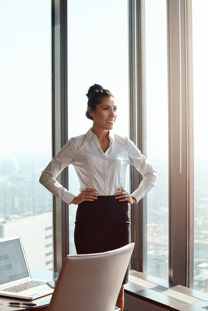 Горжусь всеми ее достижениями. привлекательная молодая деловая женщина, стоящая с руками на бедрах в офисе - Фото, изображение