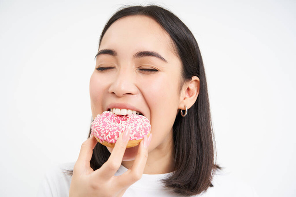 Close up of smiling pleased asian woman, eats glazed pink doughnut, enjoys eating tasty donnut, white studio background. - Fotografie, Obrázek