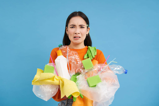 Frowning aasialainen nainen muovi roskat käsissä, valittavat ei kierrätys jätettä, seisoo pentue sinisellä pohjalla. - Valokuva, kuva