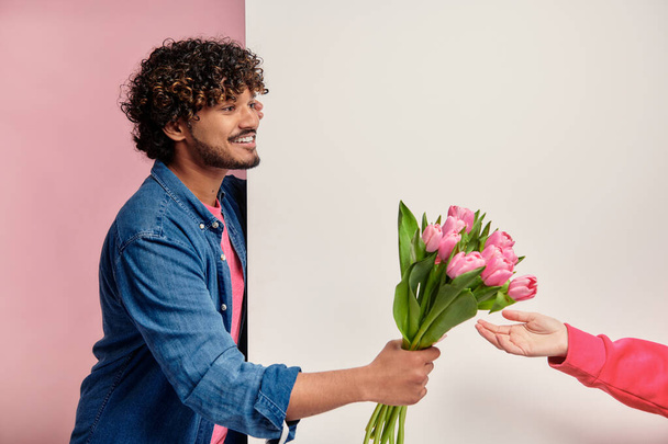 Happy Indian man giving bouquet of tulips to some woman against pink background - Photo, Image
