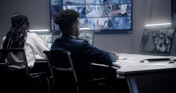 African American security operator monitors CCTV cameras with AI face recognition. Multiple big screens on the wall showing security cameras view. Colleague works at background. Social safety concept. - Photo, Image