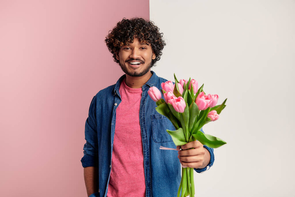 Happy Indian man stretching out bouquet of tulips against pink and white background - Photo, Image