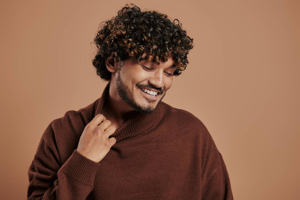 Stylish Indian man stretching sweater collar and smiling against brown background - Фото, изображение