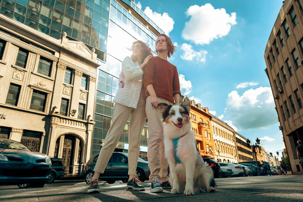 Young Caucasian couple with pet poses against background of city. Bottom view. Training purebreed aussie dog. - Foto, afbeelding