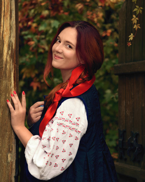 Mosolygó, vidám, fiatal, vörös hajú nő, hagyományos ukrán hímzett ruhákba öltözve, vörös szalaggal a hajában a kamerába nézve. Vintage ruha. Félhosszú portré - Fotó, kép