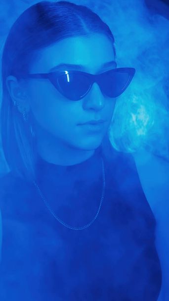 Color light people. Serious woman portrait. Neon stylish girl in trendy sunglasses concentrated focused expression on blue dark smoke background. - Fotografie, Obrázek