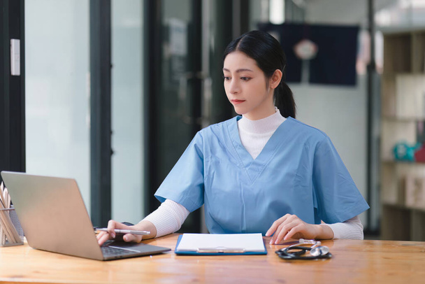 Μια συμπυκνωμένη γυναίκα γιατρός ή νοσοκόμα εργάζεται στο διαδίκτυο με ένα φορητό υπολογιστή, ενώ κάθεται σε ένα γραφείο σε ένα δωμάτιο νοσηλείας νοσοκομείου. ιατρική, τεχνολογία και υγειονομική περίθαλψη. - Φωτογραφία, εικόνα