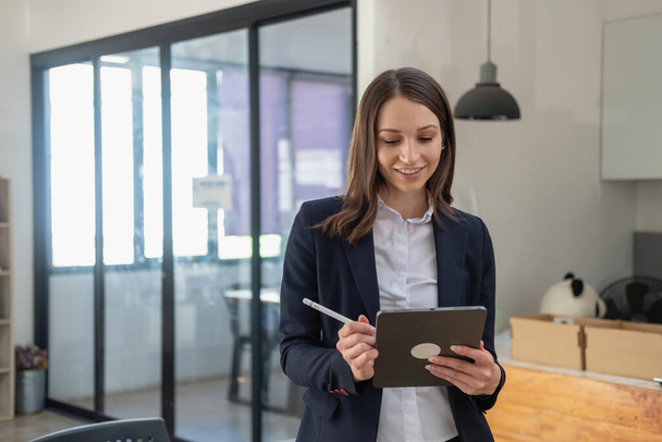 A stylish white businesswoman in a black suit standing inside the office in hands with a mobile phone, tablet, and laptop waiting for customers to contact, business people concept. - Foto, Imagem