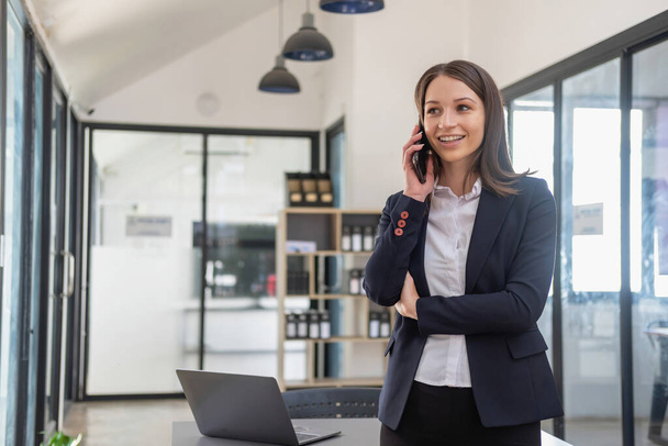 Стильная белая деловая женщина в черном костюме, стоящая в офисе в руках с мобильным телефоном, планшетом и ноутбуком в ожидании контактов с клиентами, концепция деловых людей. - Фото, изображение