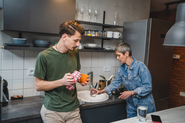 adult Caucasian man is standing in the kitchen hold a cup and cloth while his mother take care of the dishes engaged in a conversation creating a special moment of connection and bond work together - Photo, Image