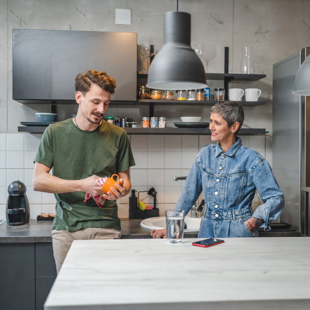大人の白人男性は台所に立っているカップと布を保持しながら、彼の母親は接続と結合の特別な瞬間を作成する会話に従事する料理を世話一緒に仕事 - 写真・画像