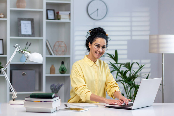 Portrait of happy successful woman at work in home office, hispanic woman smiling and looking at camera sitting at table in living room, working with laptop remotely, studying online. - Photo, image