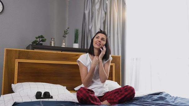 Beautiful smiling happy young woman in sleepwear talking on phone in contemporary bedroom - Фото, изображение