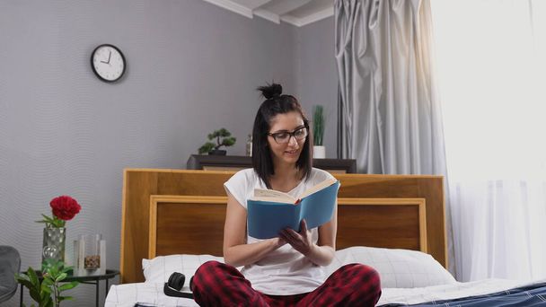 Close view of beautiful smiling carefree young brunette in glasses which reading book ,sitting on comfortable bedroom bed near big window - Fotografie, Obrázek