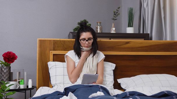 Front view of unwell sick young woman with dark hair which sitting in bed at home and using tablet pc during her flu disease - Foto, Imagem
