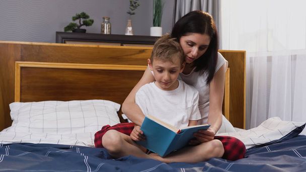 Front view of appealing satisfied loving woman and her 8-aged son which having fun together while reading book in the bedroom - Photo, Image