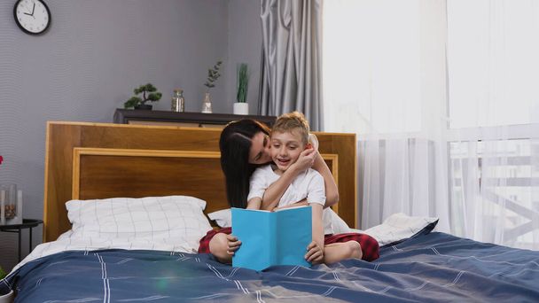 Splendide femme satisfaite de 30 ans jouant avec son beau fils de 8 ans tout en lisant un livre intéressant dans une chambre à coucher contemporaine, vue de face - Photo, image