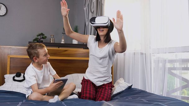 Family concept where handsome 8-aged boy using tablet pc while his happy smiling beautiful mother wearing virtual reality headset at home - Photo, image