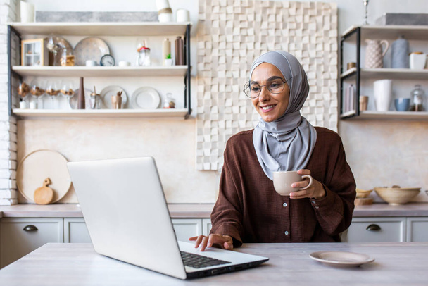 Muslim young woman in hijab sitting at the table in the kitchen at home in the morning. Drinks coffee, uses laptop, checks mail, reads news, smiles. - Foto, immagini