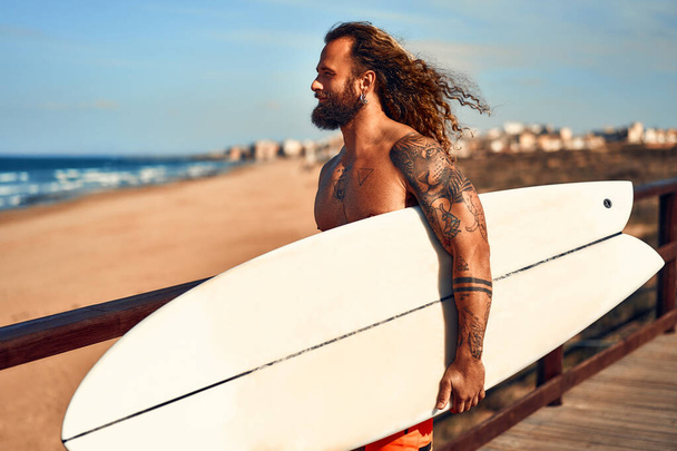 Caucasian beefy bearded man with tattoos in swimming trunks holding a surfboard walks along a wooden bridge to the sea. Sports and active recreation. - Foto, afbeelding
