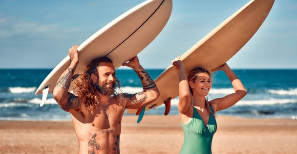 ビーチでサーファーのカップル。水着姿の白人女性と海の近くのサーフボードで水着姿の髭のバフマン。スポーツとアクティブなレクリエーション. - 写真・画像