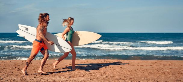 A couple of surfers on the beach. Caucasian woman in a swimsuit and a bearded buff man with tattoos in swimming trunks with surfboards near the sea. Sports and active recreation. - Photo, Image