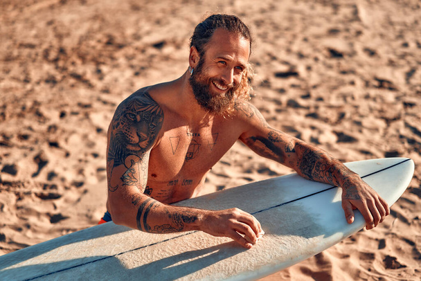 Homme barbu caucasien costaud avec des tatouages dans les maillots de bain cire une planche de surf près de la mer. Sports et loisirs actifs. - Photo, image
