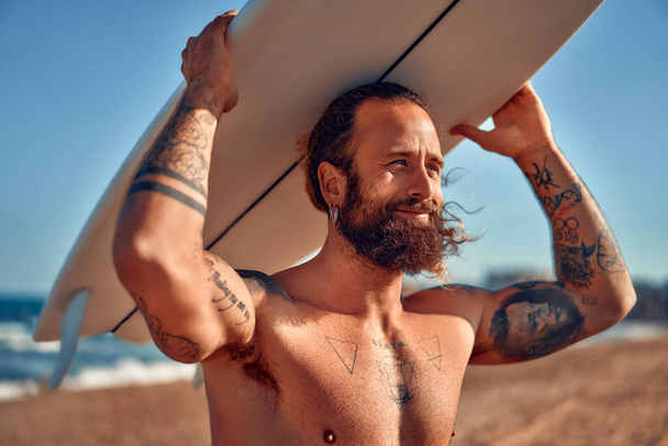 Běloch masitý surfař vousatý muž s tetováním a dlouhými vlasy v plaveckých trenýrkách se surfařem u moře. Sportovní a aktivní rekreace. - Fotografie, Obrázek