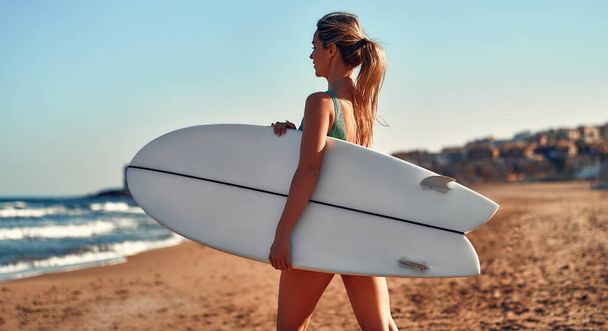 海の近くの砂浜にサーフボードが立っている水着で美しい白人女性。スポーツとアクティブなレクリエーション. - 写真・画像