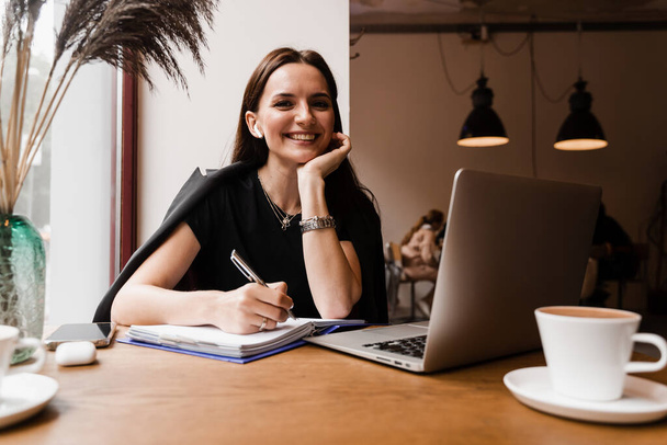 Suorapuheinen tyttö, jolla on kannettava tietokone, istuu kahvilassa, hymyilee ja iloitsee onnistuneesta työstä IT-yrityksessä. Iloinen nuori nainen ohjelmoija toimii etänä kannettavalla tietokoneella ja yrittää noudattaa määräaikaa - Valokuva, kuva