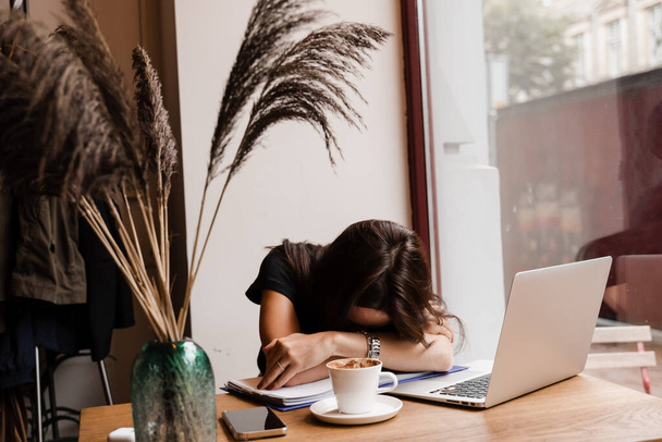 Κοιμωμένη με λάπτοπ στο τραπέζι του καφέ. Κουρασμένη νεαρή γυναίκα με κλειστά μάτια έχουν ένα διάλειμμα στη δουλειά και τον ύπνο και χαλάρωση στο φορητό υπολογιστή στο χώρο εργασίας λόγω της υπερωριακής εργασίας - Φωτογραφία, εικόνα