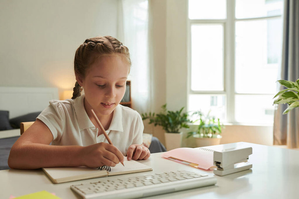 Χαμογελώντας preteen κορίτσι κάνει την εργασία στο γραφείο της, γράφοντας στο βιβλίο - Φωτογραφία, εικόνα