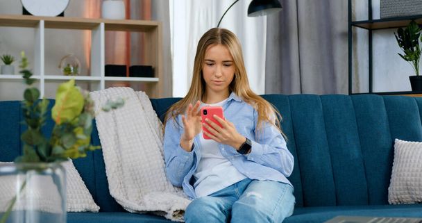 Portrait of attractive in good mood relaxed young girl with light long hair in casual clothes resting at home on comfortable sofa and using her smartphone - Foto, Imagen