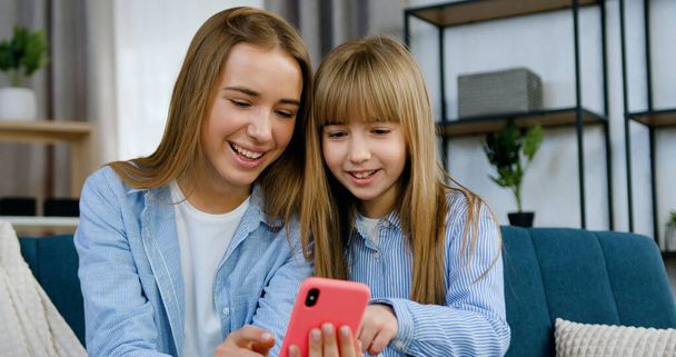 Attractive smiling friendly two 20-aged and 10-aged sisters sitting on soft couch at home and watching funny videos on smartphone while spending joint free time - Foto, afbeelding