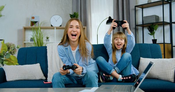 Lovely friendly cheerful two different ages girls playing video games using joysticks and celebrating victory with raised hands when spending joint free time at home - Φωτογραφία, εικόνα