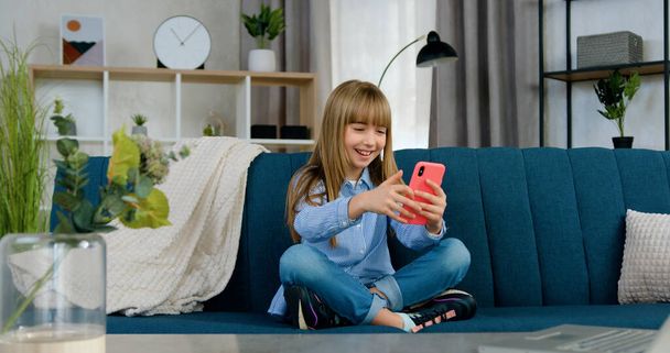 Portrait of good-looking cheerful happy 10-aged girl which sitting in lotus pose on soft couch at home and playing with smartphone revisioning funny videos - Fotografie, Obrázek
