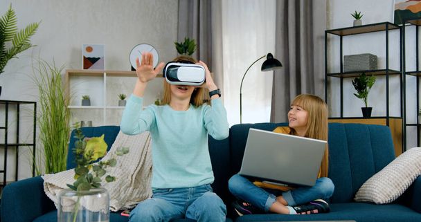 Likable smiling modern light haired 20-aged girl uses virtual reality headset near her younger sister which applying computer,joint leisure at home concept - Photo, image