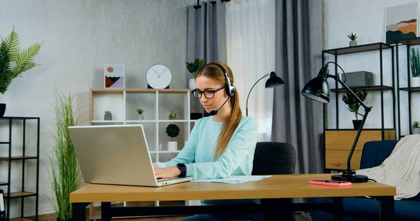 Likable purposeful smart modern female student in headset wears glasses dressed in casual clothes working on laptop during isolation at home,remote education concept - Fotografie, Obrázek