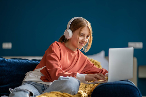 Positiv lächelndes Teenie-Mädchen mit Kopfhörern, das Videofilme auf dem Laptop anschaut, wie es zu Hause auf der Couch sitzt. Freizeit, Erholung, Unterhaltung Zeitvertreib der Teenager Wahl Film, Zeichentrick auf dem Computer zu sehen.  - Foto, Bild