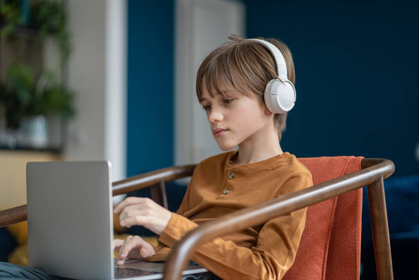 Serious attentive caucasian teen boy in headphones having online class on laptop sitting on chair in living room home. Remote, distance education, homeschooling. Listening lection, lesson, webinar. - Photo, Image