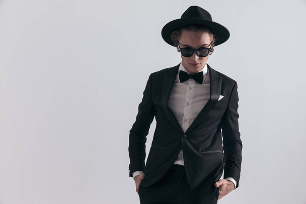elegant fashion groom with hat and sunglasses posing with hands in pockets in front of grey background in studio - Foto, Imagen