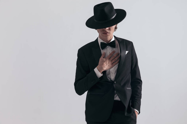 jovem elegante em smoking preto com chapéu nos olhos segurando uma mão no bolso e tocando no peito na frente de fundo cinza em estúdio - Foto, Imagem