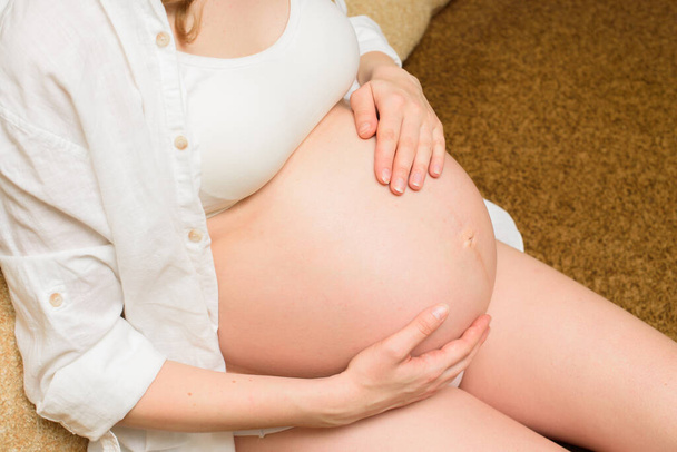 Těhotná dívka hladí své břicho, sedí na gauči. Těhotná žena drží břicho v ruce. Matka čeká dítě - Fotografie, Obrázek