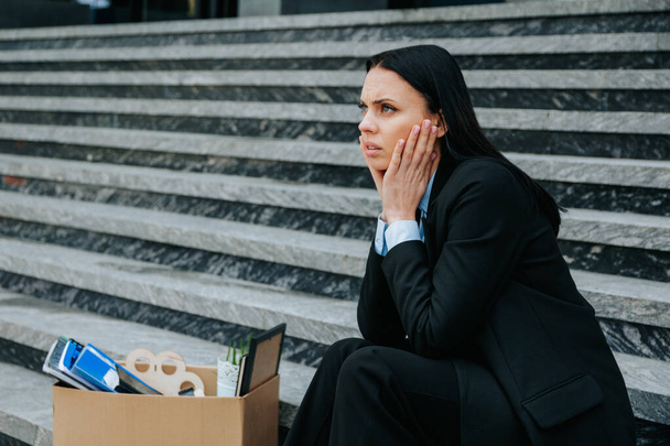 woman sitting on stairs, symbolizing the struggle of job loss and worklessness due to dismissal and unemployment. a woman sitting on stairs, cardboard box beside her - Фото, изображение