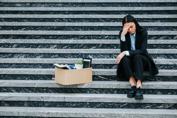 A photo of a woman sitting on stairs, cardboard box beside her, as she comes to terms with the reality of her fired job and workloss. - Foto, Imagen