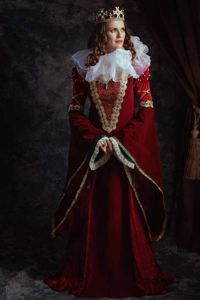 Full length portrait of pensive medieval queen in red dress with white collar and crown on dark gray background. - Фото, зображення