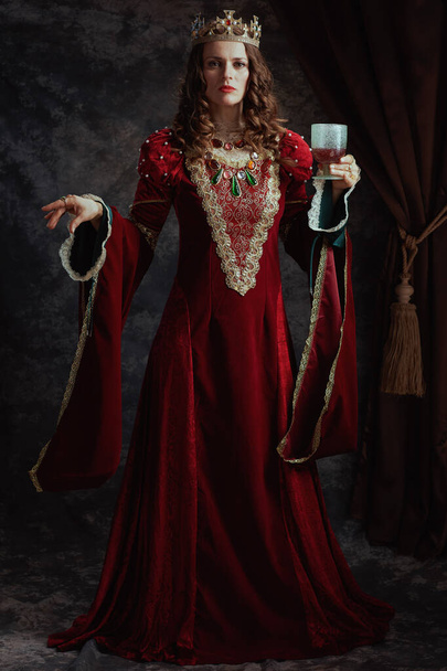 Full length portrait of medieval queen in red dress with goblet and crown on dark gray background. - Фото, зображення