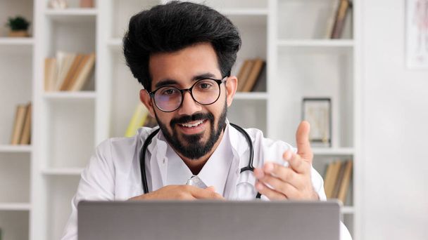 Smart friendly male Indian clinician in white coat using laptop for remote patient consultation in hospital office. Telemedicine. Online appointment with a doctor - Photo, image