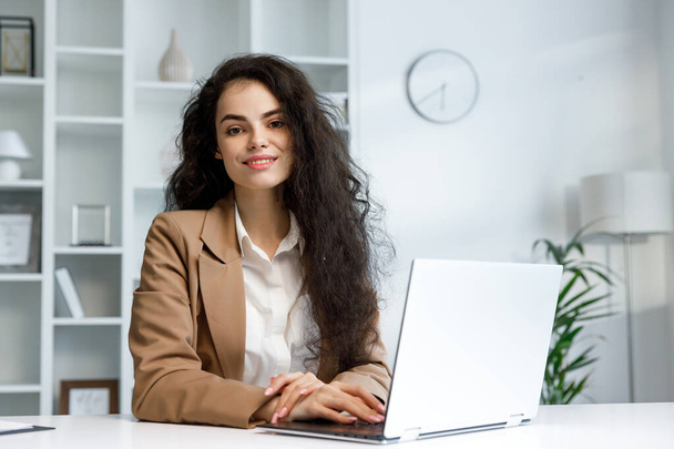 Young curly brunette girl with a laptop looks at the camera. Freelancer portrait working from home, remote work. Successful manager girl, in a white office. Reports online - Zdjęcie, obraz
